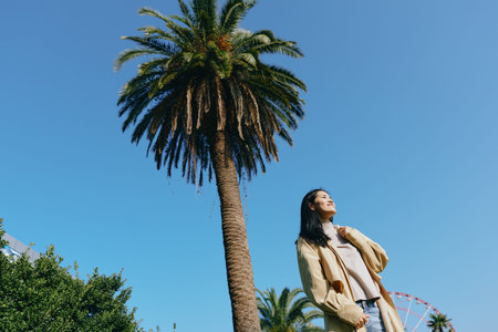 Woman in casual outfit standing beside a tall palm tree under a clear blue sky in a sunny park, an outdoor scene showcasing nature, leisure and relaxed lifestyle in bright lightの写真素材
