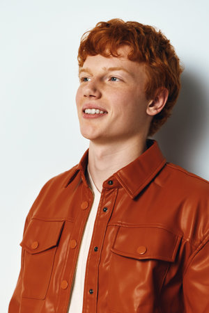 Young man with red hair wearing brown leather jacket smiling and looking aside with white background for portrait and casual fashion style.の写真素材