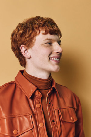 Young man with red hair and freckles smiling in brown leather jacket and turtleneck sweater against beige background in studio portrait.の写真素材