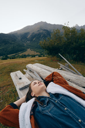 woman lying on a wooden bench in a meadow with mountains in the background, portrait of relaxation outdoors in countryside, casual denim jacket and shearling coat, peaceful scenic escapeの写真素材