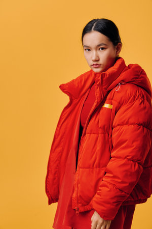 Young woman with long dark hair wearing a red puffer jacket and matching red dress standing against a solid orange background looking at camera with serious expression and side pose.の写真素材