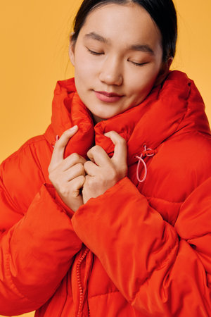 Young woman with closed eyes wearing bright orange puffer jacket on yellow background. Cozy winter clothing, warmth, comfort and casual style with peaceful expression and soft lighting.の写真素材