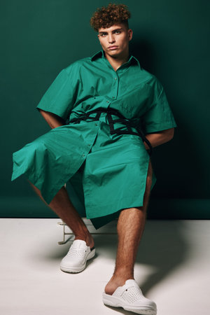 Young man with curly hair wearing green oversized shirt dress and white slip-on shoes sitting against dark green background in studio with casual modern fashion style.の写真素材