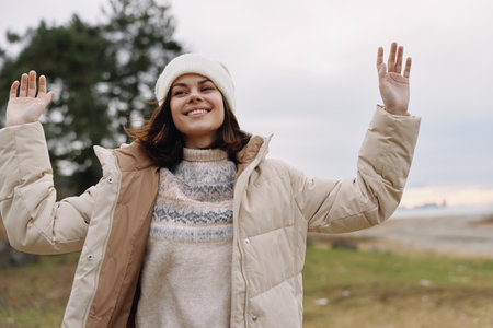 Woman in a warm winter coat outdoors, arms raised in joy, smiling at the camera against a cloudy sky...