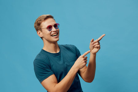 smiling caucasian man wear sunglasses turquoise t-shirt point finger blue background cheerful positive emotion casual style summer fashion conceptの写真素材