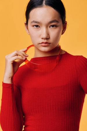 Young woman with serious expression wearing red sweater holding red sunglasses posing on yellow...
