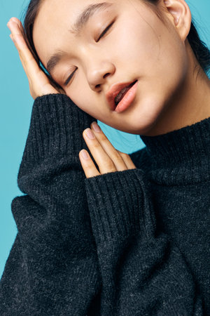 Woman with closed eyes and relaxed face wearing dark sweater posing with hand on cheek against blue background. Portrait of calm peaceful female with smooth skin and natural expression.の写真素材
