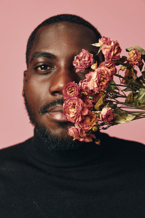 African man with beard and black turtleneck holding dried pink roses near face on pink background in creative conceptual visual style portrait with floral details and emotion.の写真素材