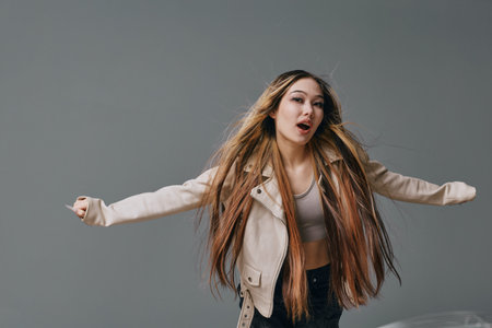 Young woman with long hair wearing casual jacket and crop top stretching arms with opened mouth on gray background. Fashion, studio portrait, beauty and confidence concept.の写真素材