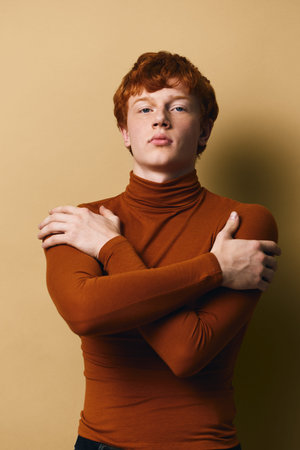 Young man with red hair wearing brown turtleneck posing with crossed arms against beige background. Serious expression, studio portrait with soft lighting and minimalistic style.の写真素材
