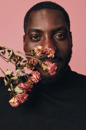 African man with dark skin and beard holding dried pink flowers near face in creative conceptual visual style on pink background. Portrait with artistic expression and floral detail.の写真素材