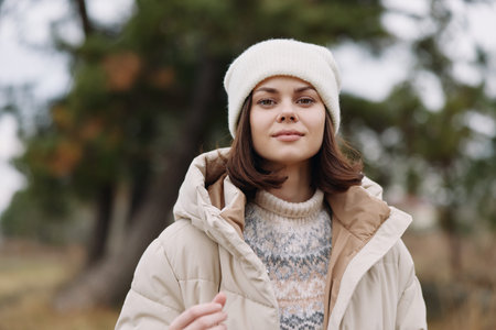 Winter portrait of a woman in a beige coat and knit sweater, wearing a white beanie, standing...