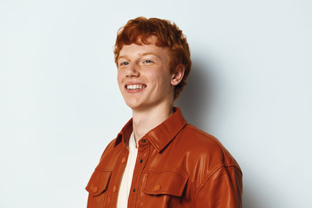 Young man with red hair smiling confidently against a white background. Casual style wearing a brown...