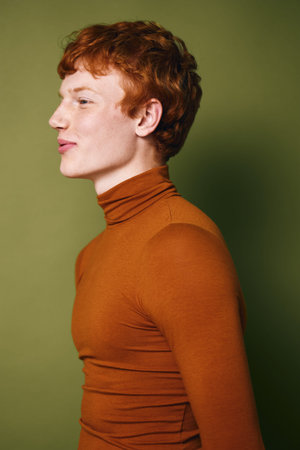 Young man with red hair and pale skin wearing orange turtleneck sweater posing in profile against green background. Stylish casual portrait in studio with neutral expression and soft lighting.の写真素材