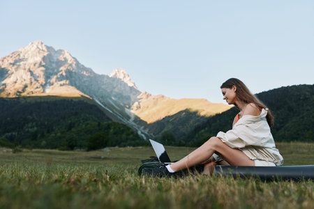 A relaxed woman sits on a grassy field with a laptop, enjoying nature and work balance in a peaceful outdoor office scene, framed by distant mountains and clear sky.の写真素材