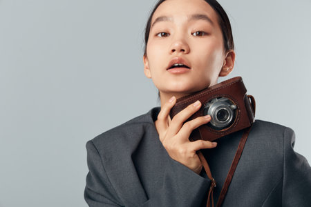 Young woman holding vintage camera close to face wearing oversized dark jacket posing against plain...