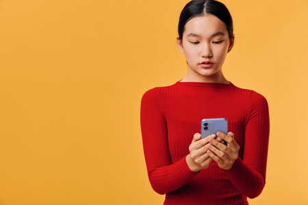 Young woman in red sweater holds smartphone and uses touch screen on orange background. Casual Asian...