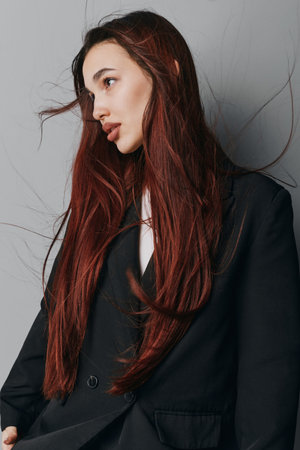 Woman with long red hair wearing black blazer posing against gray wall. Fashion model with natural...
