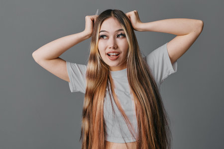 Young woman with long hair wearing casual shirt posing against gray background with playful expression and hands on head for lifestyle, emotion and portrait photography.の写真素材