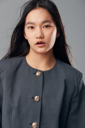 Portrait of Asian woman with long dark hair wearing gray blazer with decorative buttons on gray background. Young female model looking at camera with natural expression in studio light.の写真素材