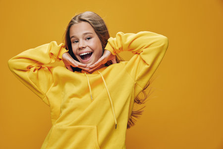 Girl smiling and posing with hands under chin wearing yellow hoodie with long hair on yellow...