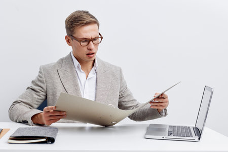 Professional young man in business attire working at desk with laptop and documents, looking confused or surprised while reviewing paperwork in bright modern office. Business career conceptの写真素材