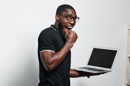 African American man with dark skin and glasses holding laptop and biting fist in excitement or...