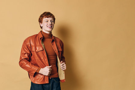 Young man with red hair wearing orange leather jacket and brown turtleneck posing smiling on beige background in casual style with hands in pockets.の写真素材