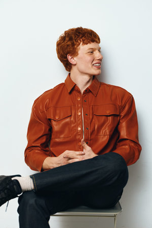 Young man with red hair sitting on chair looking sideways smiling, wearing brown leather shirt and black pants in casual studio portrait on white background.の写真素材