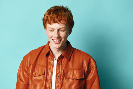 Young man with red hair wearing brown leather jacket smiling with eyes closed against blue background. Portrait of cheerful male adult with casual style isolated indoors.の写真素材