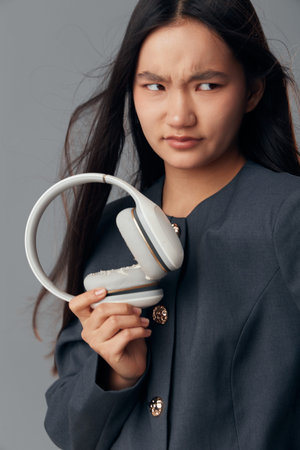 Woman holding broken wireless headphones with confused expression. Asian female in dark jacket examining damaged audio device in studio with neutral background. Technology and lifestyle concept.の写真素材