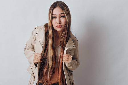 Young woman with long brown hair wearing beige jacket posing confidently against white background. Casual fashion style with natural makeup and relaxed expression in studio setting.の写真素材