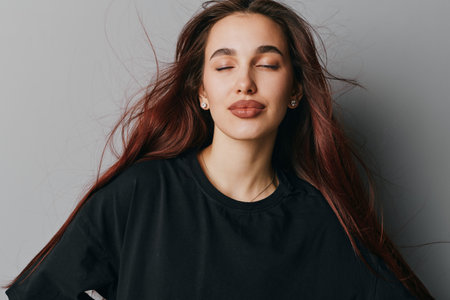 Woman with long hair and closed eyes wearing black shirt posing against gray background for beauty, relaxation, and calm expression in studio portrait with natural makeup and earrings.の写真素材