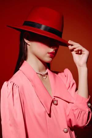 Fashionable woman in pink jacket and red hat tilts hat visor with hand, lips painted red. Creative...