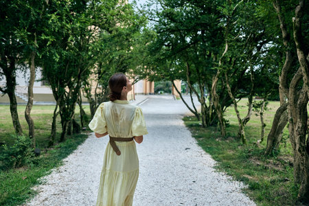 Woman in a pale dress walks along a gravel path under lush trees in a peaceful park, conveying calm, exploration and a timeless outdoor momentの写真素材