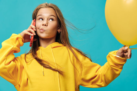 Girl in yellow hoodie holding yellow balloon and talking on red phone with playful expression on...