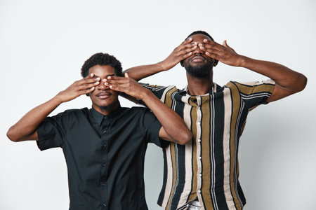 Two African men with dark skin covering their eyes with hands standing against white background....