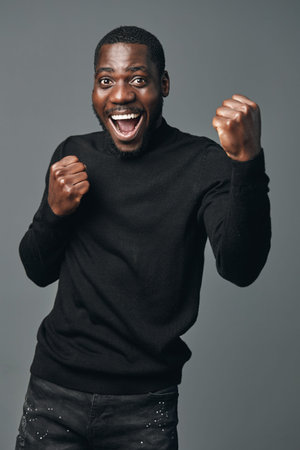 African man with dark skin wearing black sweater showing excitement and joy with clenched fists in creative conceptual visual style on gray background. Happy, energetic and expressive portrait.の写真素材