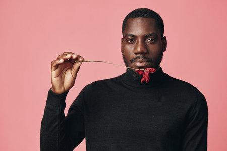 Creative conceptual portrait of African man in black turtleneck holding red chili pepper near mouth on pink background. Visual style with focus on expression and color contrast.の写真素材
