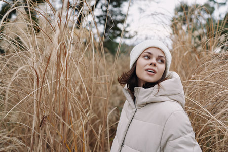 Woman in a light winter coat stands among tall dried grasses, gazing upward in a chilly outdoor...