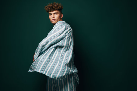 Young man with curly hair wearing striped shirt and pants posing against dark green background in studio for fashion portrait and style concept.の写真素材
