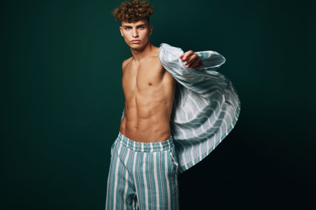 Young man with curly hair wearing striped pajamas shirt open showing toned chest and abs on dark...