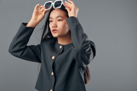 Woman posing with sunglasses in hands against gray background wearing modern black jacket with buttons. Young female model with long hair styling her glasses for casual fashion shoot or portrait.の写真素材
