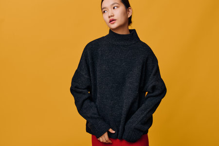 Young woman wearing oversized black sweater and red skirt posing on yellow background looking aside with thoughtful expression in studio for fashion portrait concept.の写真素材