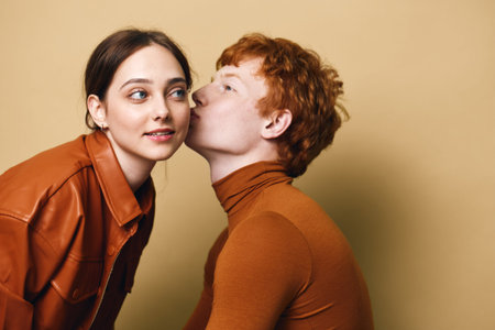 Man and woman in brown clothing, with man kissing woman on cheek against beige background. Couple showing affection, red hair and casual style with intimate moment.の写真素材
