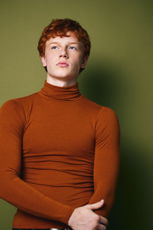 Young man with red hair wearing orange turtleneck sweater posing against green background in studio. Portrait of confident male model with crossed hands looking away thoughtfully indoors.の写真素材
