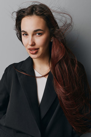 Woman with long hair and natural makeup wearing black blazer posing against gray background with confident expression and wind blowing through hair in studio portrait.の写真素材