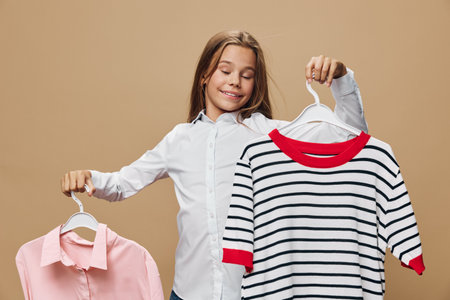 Girl holding two clothes hangers with casual tops smiling in studio, choosing outfit, comparing fashion styles, clothing, youth, lifestyle and wardrobe selection concept.の写真素材