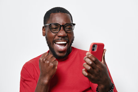 African American man with dark skin wearing glasses and red shirt smiling excitedly while looking at smartphone, celebrating success or good news indoors on white background.の写真素材