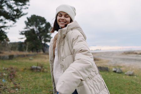 Woman in a warm beige coat smiles outdoors in a windy countryside, wearing a knit hat and cozy...